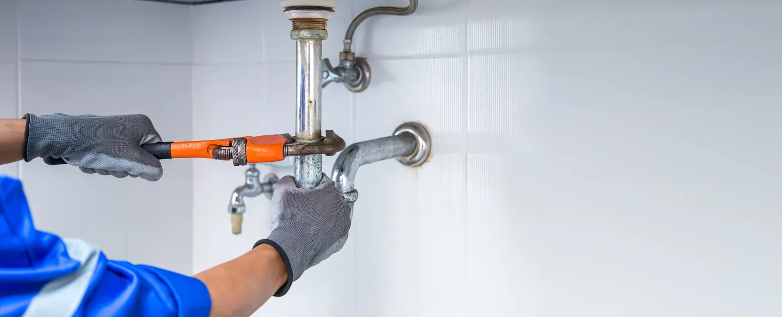 Plumber fixing a pipe under the sink using a wrench wearing protective gloves and blue uniform.