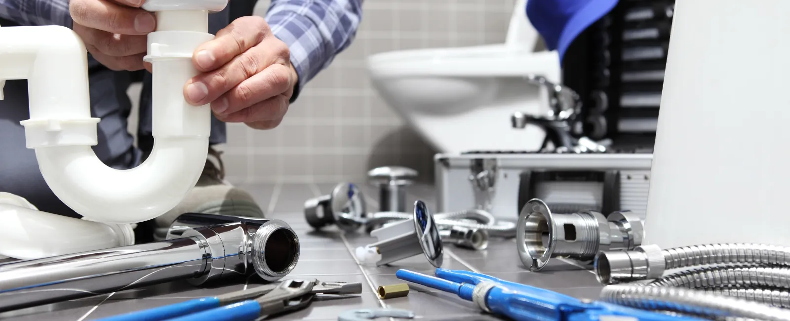 Plumber installing white PVC pipe under bathroom sink surrounded by tools and metal plumbing parts on tiled floor