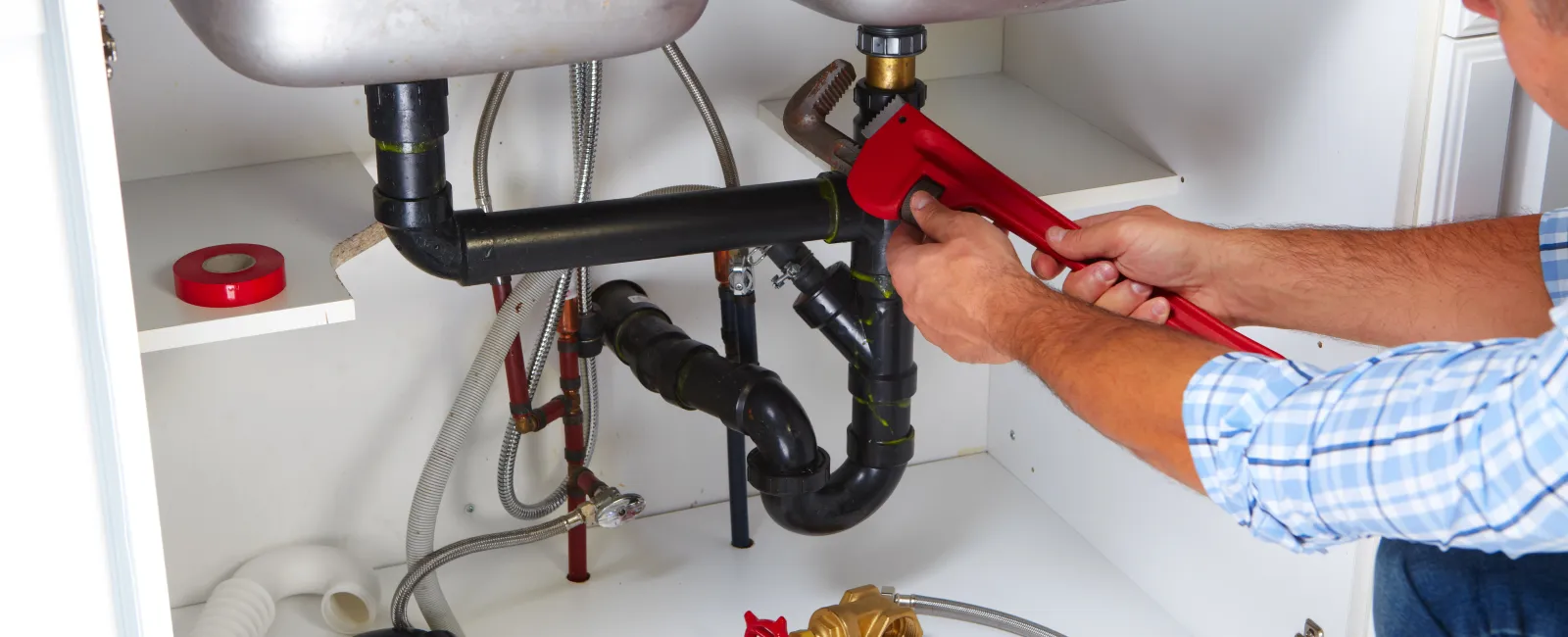 Plumber using a red wrench to fix black sink pipes under kitchen cabinet with tools on white shelf