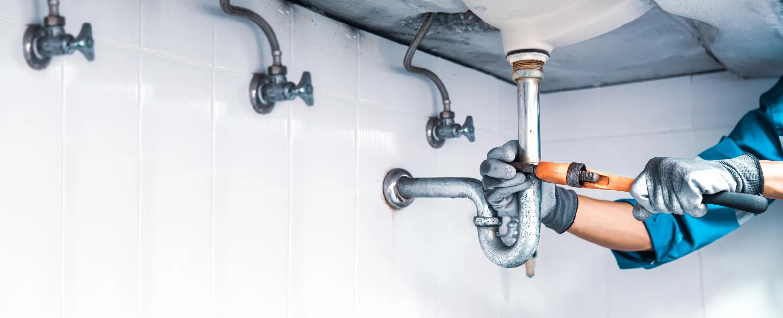 Plumber wearing gloves repairing a metal pipe underneath a sink in a tiled bathroom or kitchen setting.