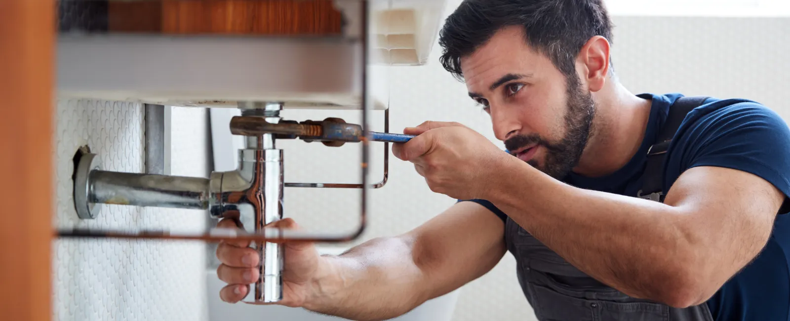 Man wearing overalls fixing bathroom sink plumbing with a wrench under a white basin in natural light.