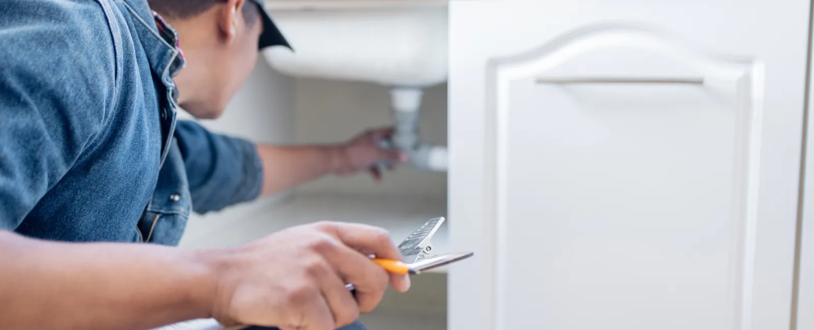 Plumber inspecting and repairing white kitchen cabinet plumbing with adjustable wrench in hand.