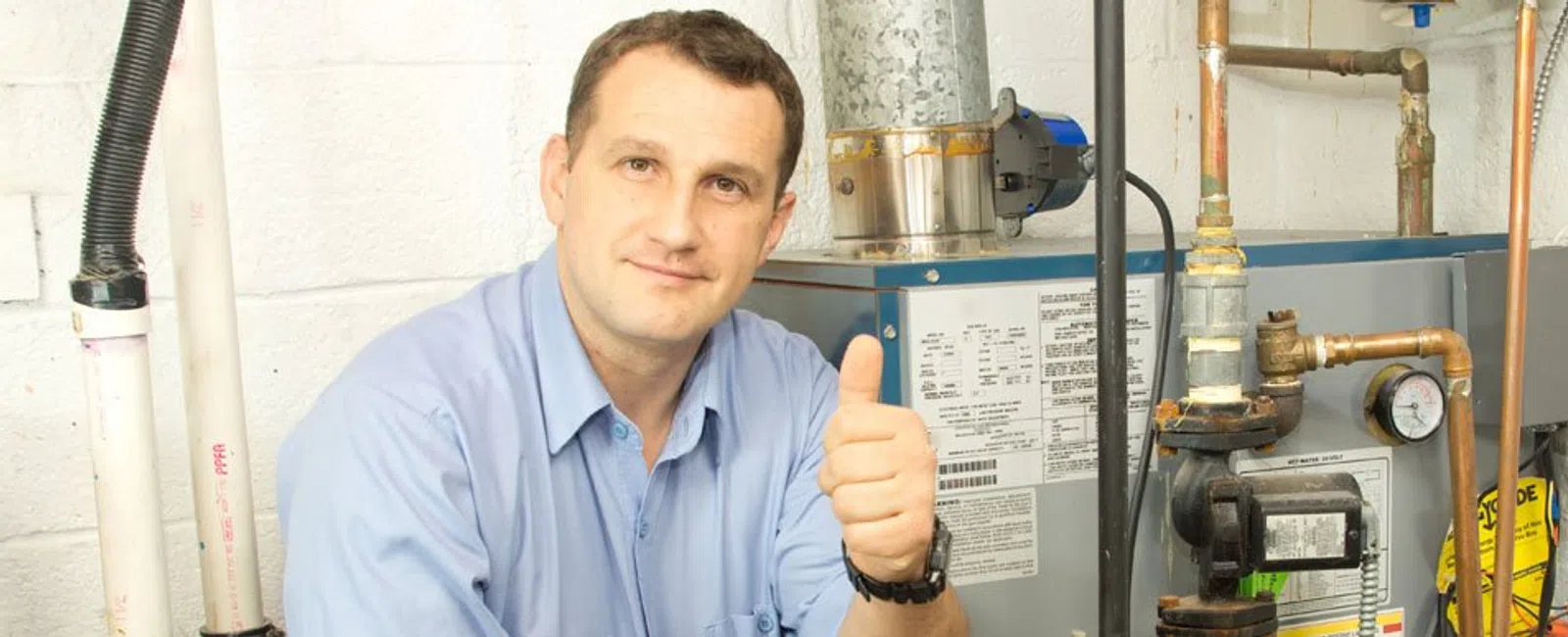 Technician giving thumbs up next to heating system and pipes in a utility room or basement.