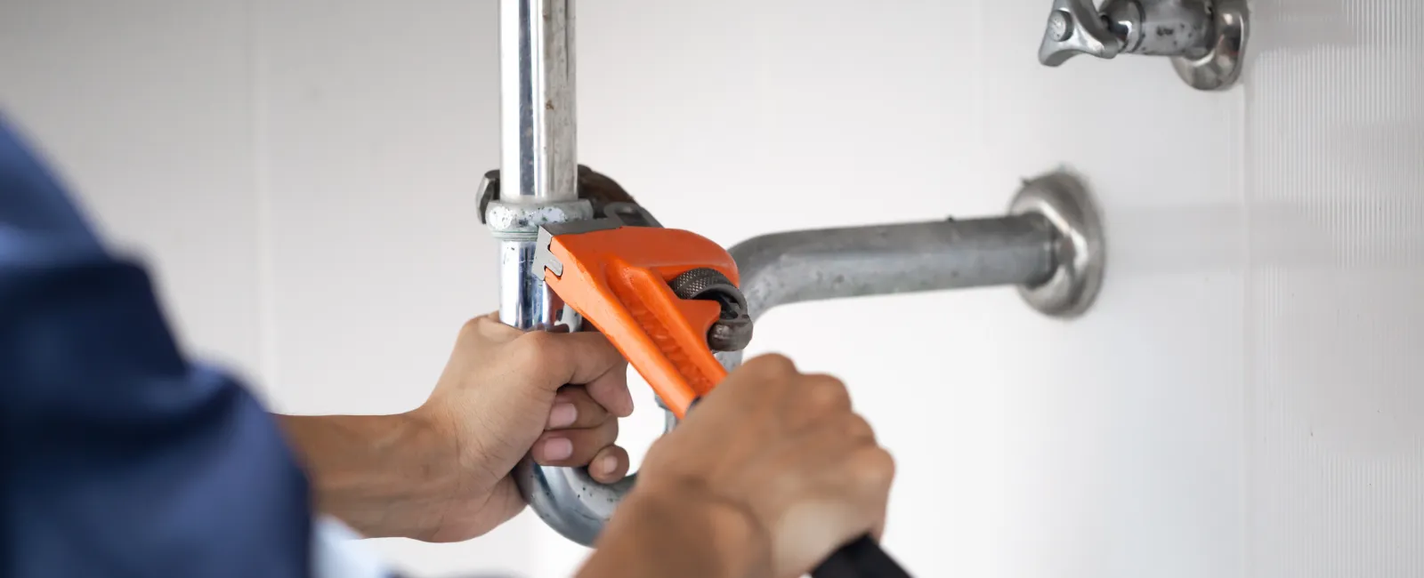 Plumber using an orange pipe wrench to fix chrome pipes under a white tiled sink area.