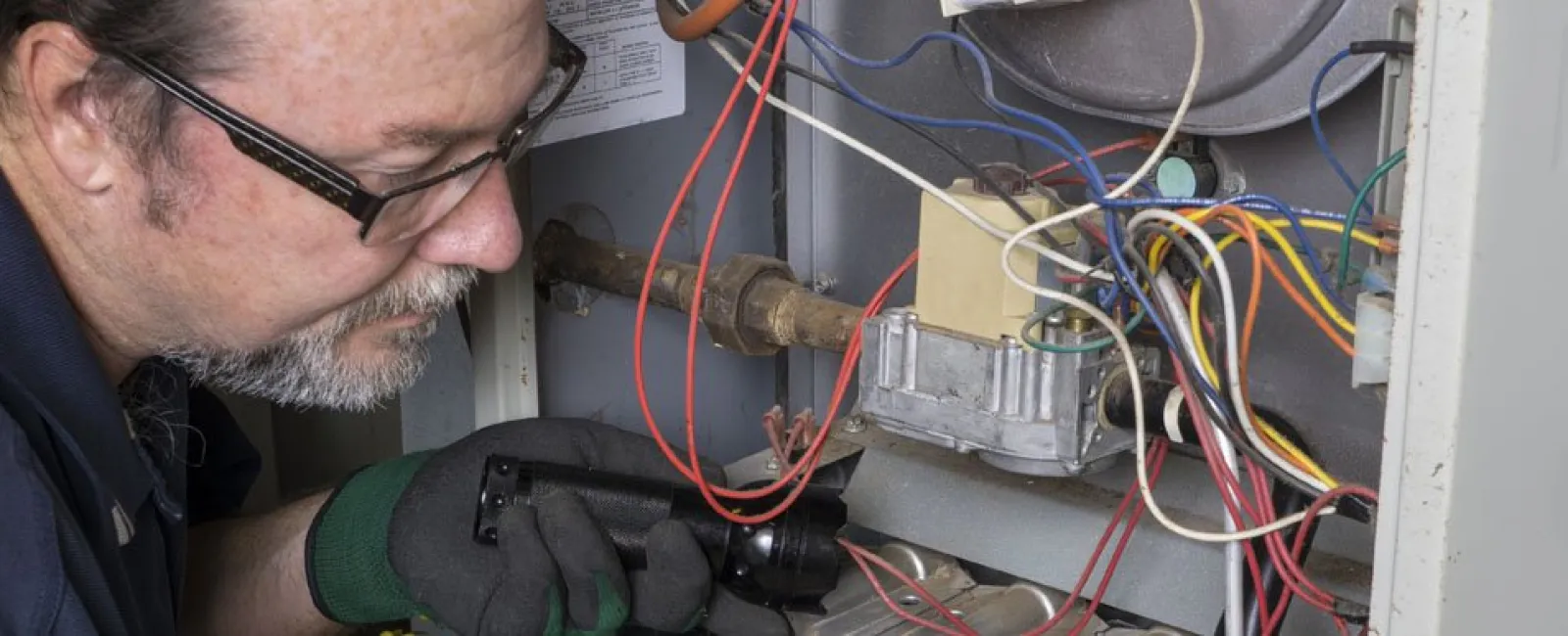 Technician repairing a furnace with colorful wires and electrical components inside a metal casing.