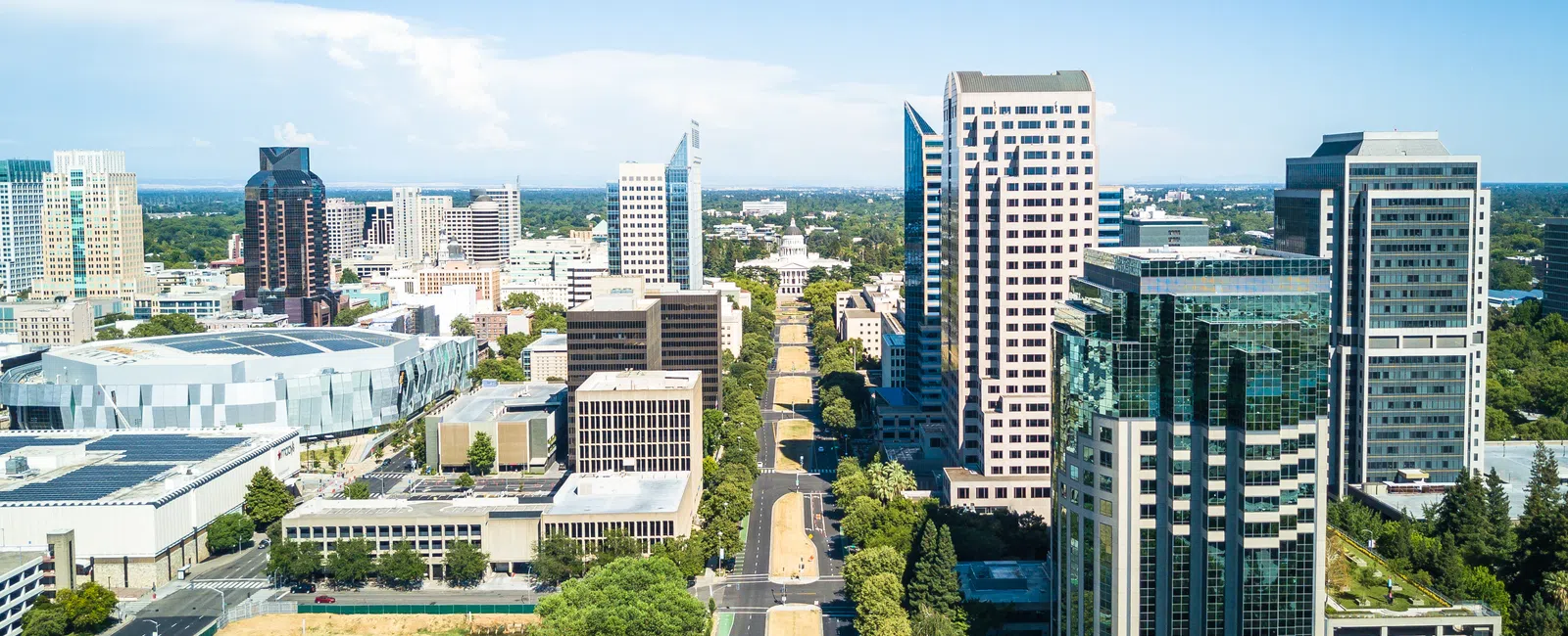 Aerial view of Sacramento downtown with high-rise buildings, a wide boulevard, and green trees under a clear blue sky.