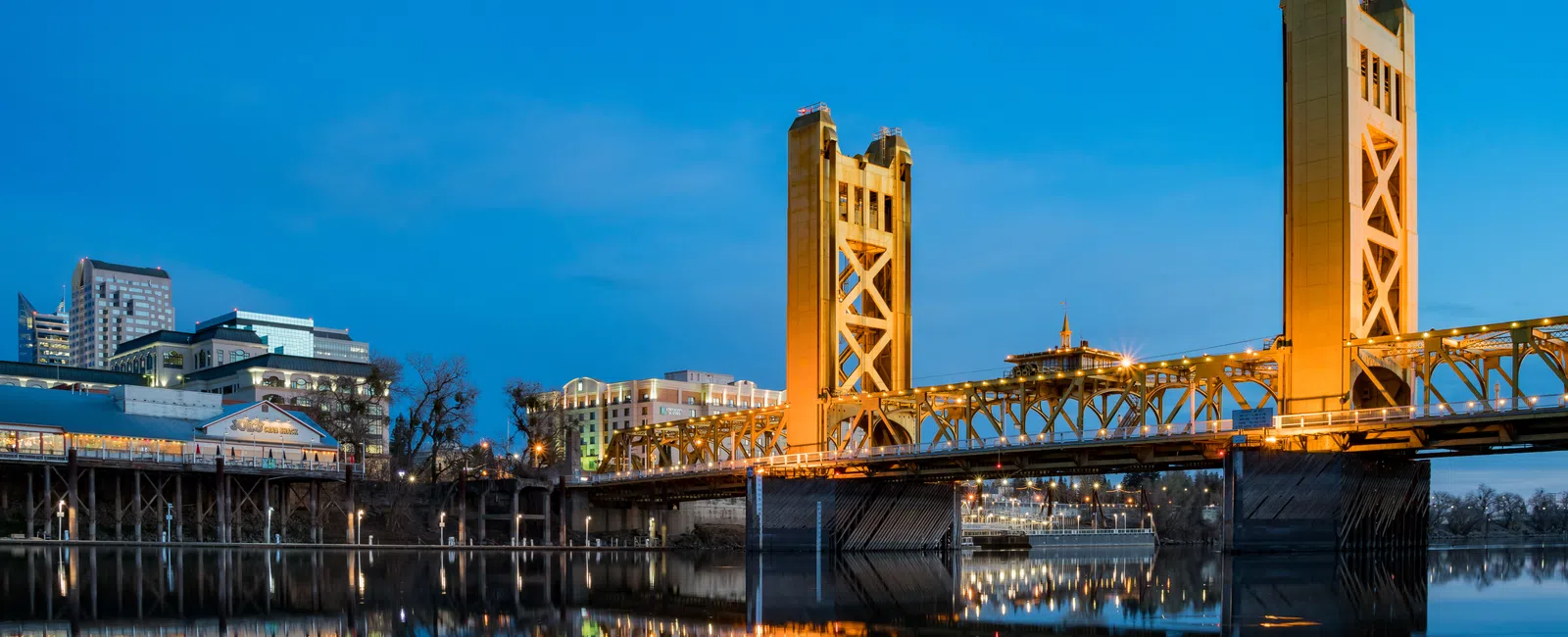 Golden Tower Bridge illuminated at dusk reflected in calm river water with city skyline in background.