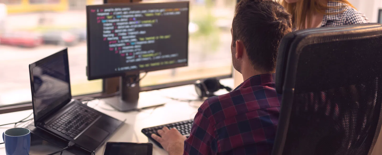 Two software developers collaborate at a desk with code on a monitor and a laptop in a modern office.