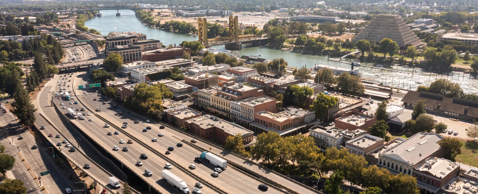Aerial view of a busy highway, river, bridge, and historic buildings in a sunny urban area with green trees.