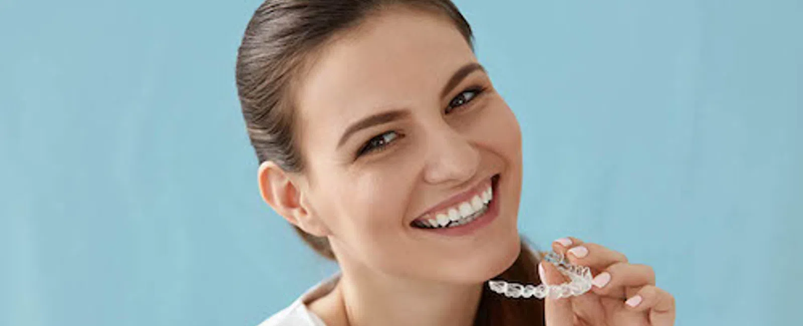 Smiling woman holding a clear dental aligner against a blue background promoting orthodontic care.