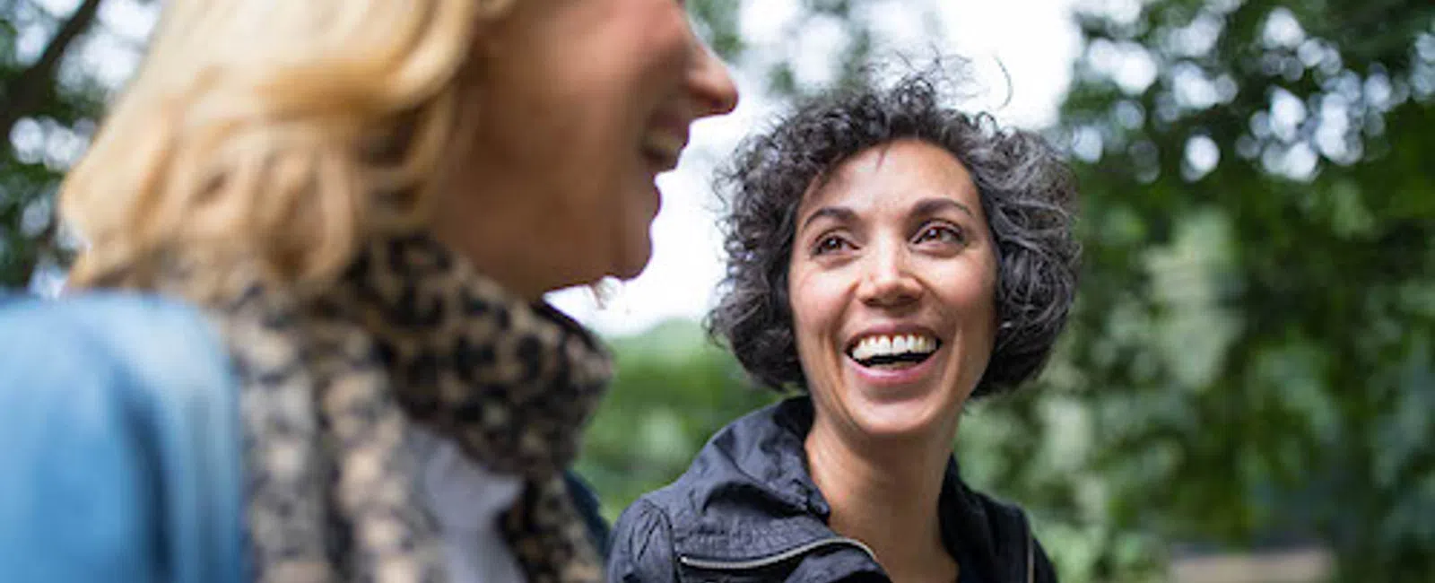 Two women smiling and talking outdoors near a lake with greenery in the background on a cloudy day