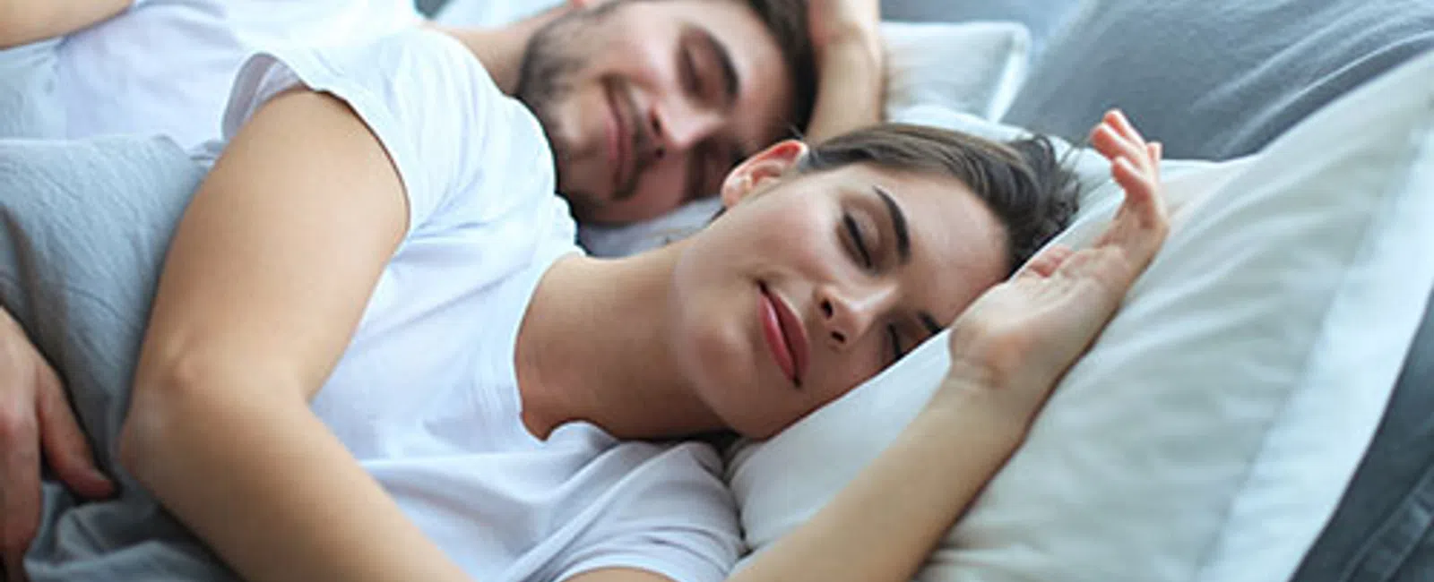 Young couple peacefully sleeping together in bed with white and gray bedding, showing comfort and relaxation.