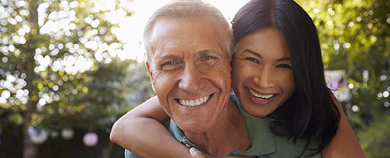 Smiling elderly man getting a piggyback hug from a cheerful younger woman outdoors in a sunny garden.