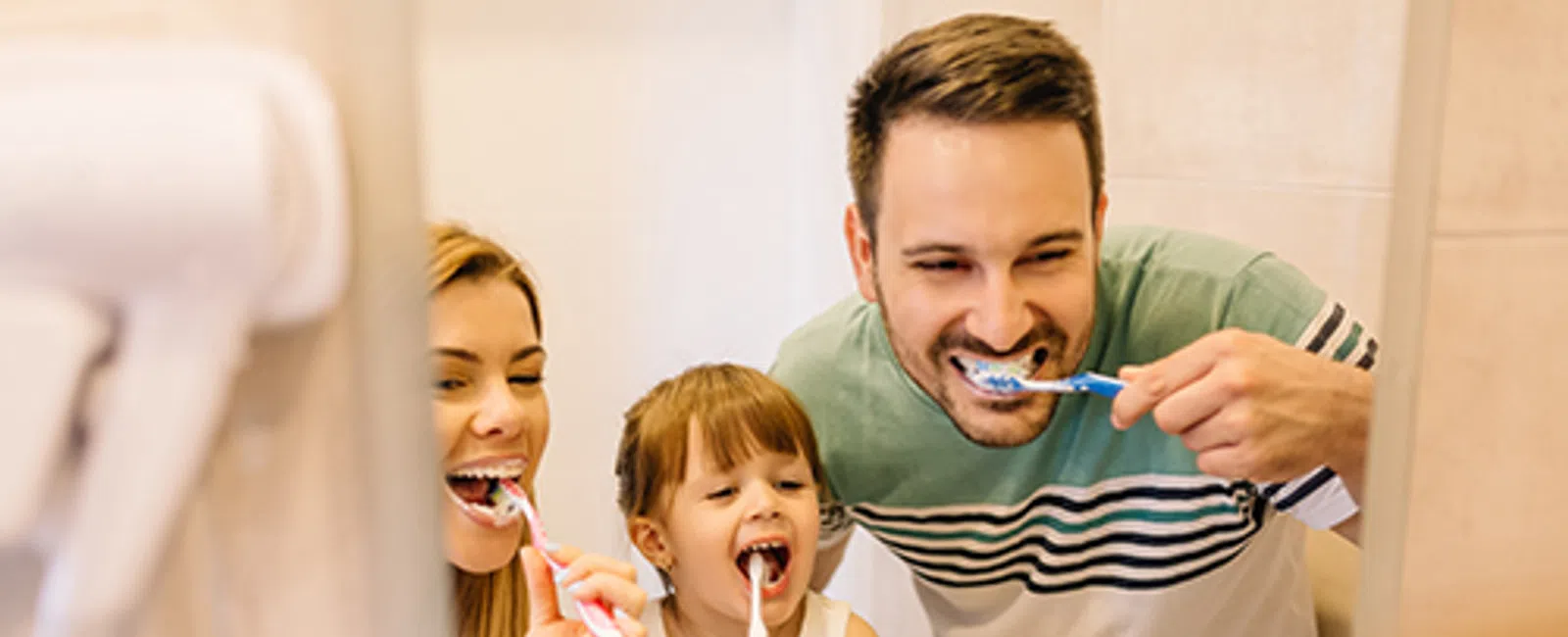 Family of three brushing their teeth together in front of a bathroom mirror, smiling and enjoying oral care.