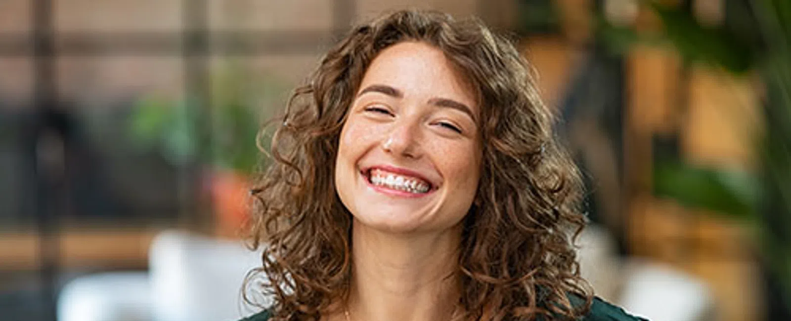 Smiling young woman with curly hair wearing green blouse and gold necklace in cozy indoor setting.