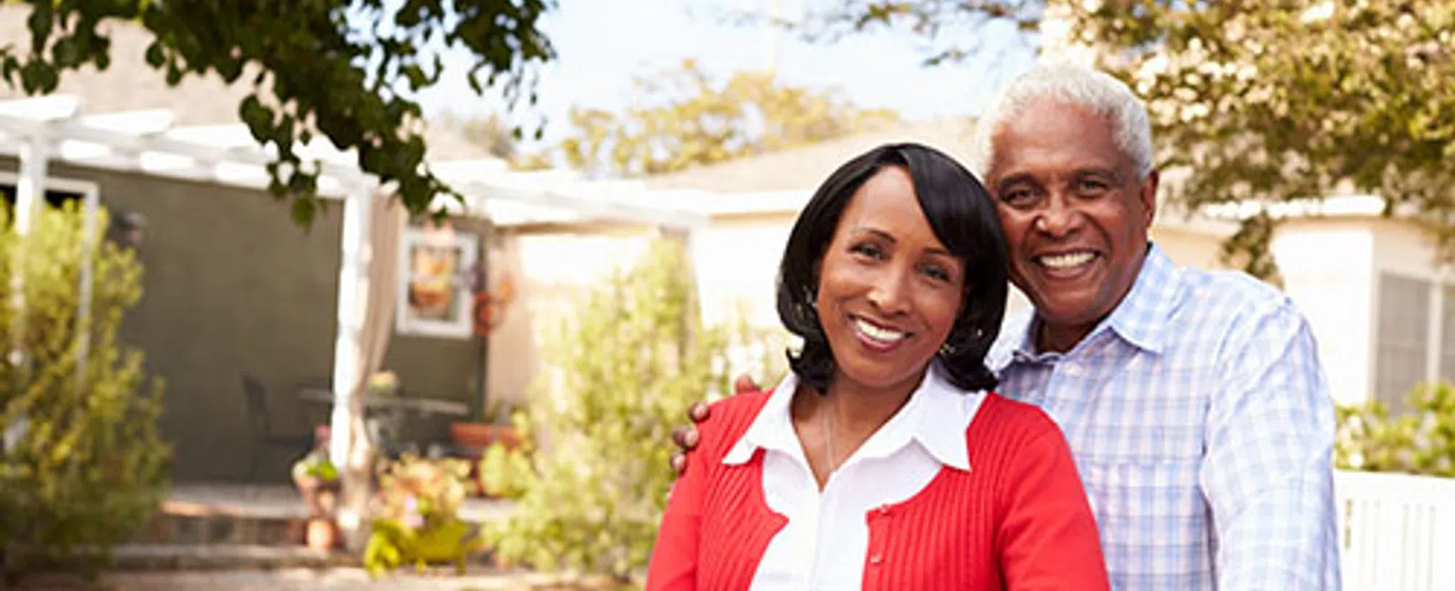 Smiling mature African American couple outdoors in their backyard on a sunny day, posing by a white fence.