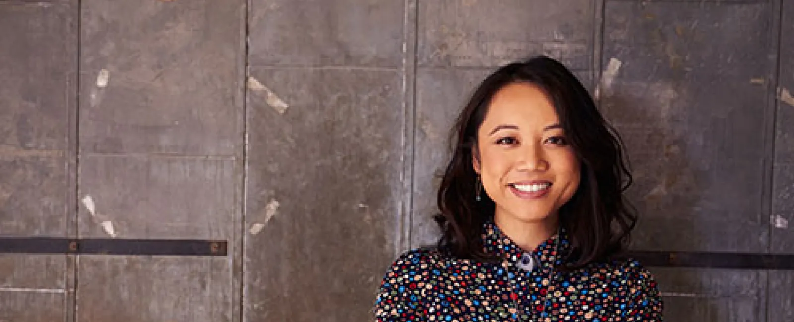 Confident woman with crossed arms wearing a colorful polka dot shirt standing against a rustic metal wall.