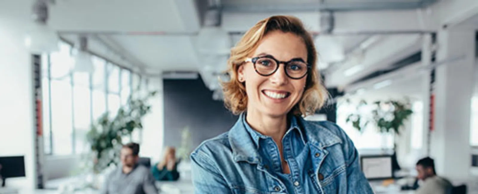 Smiling woman in glasses and denim jacket crossing arms in modern office with coworkers and computers in background
