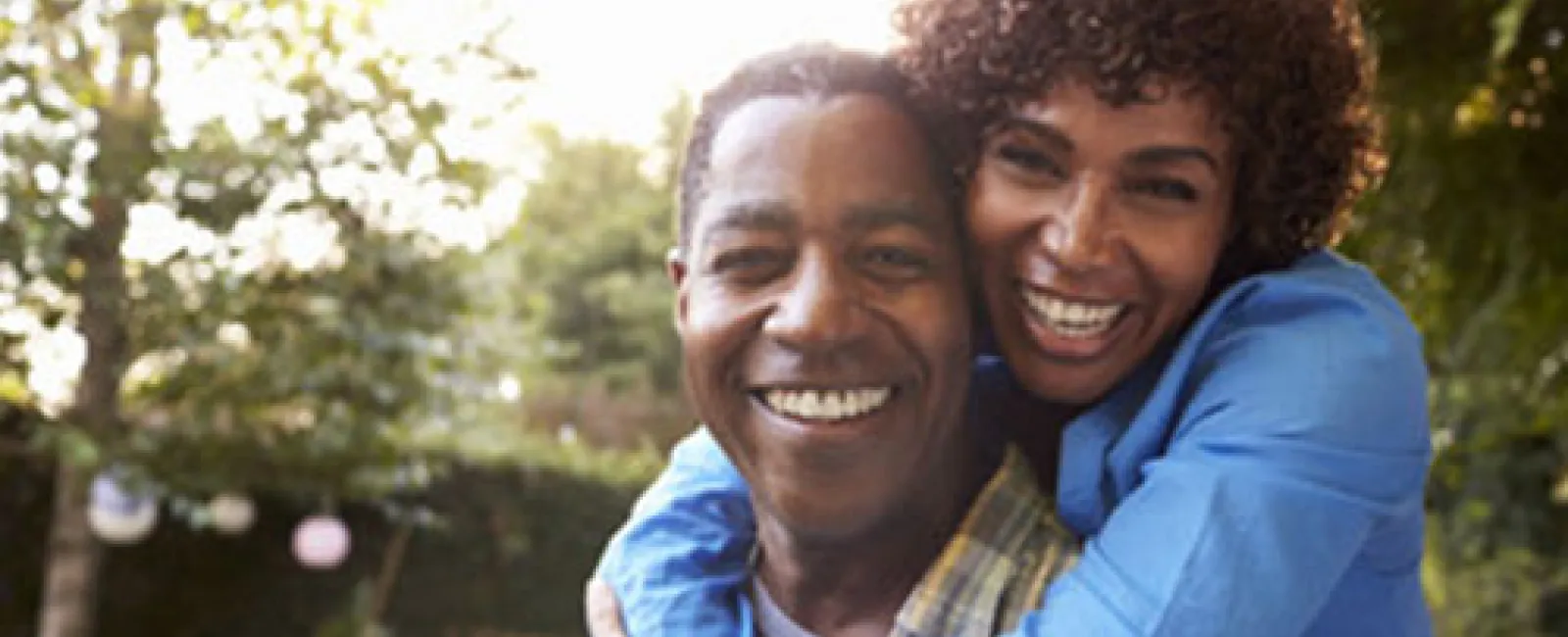 Happy couple outdoors smiling as woman hugs man from behind in a sunny garden setting.