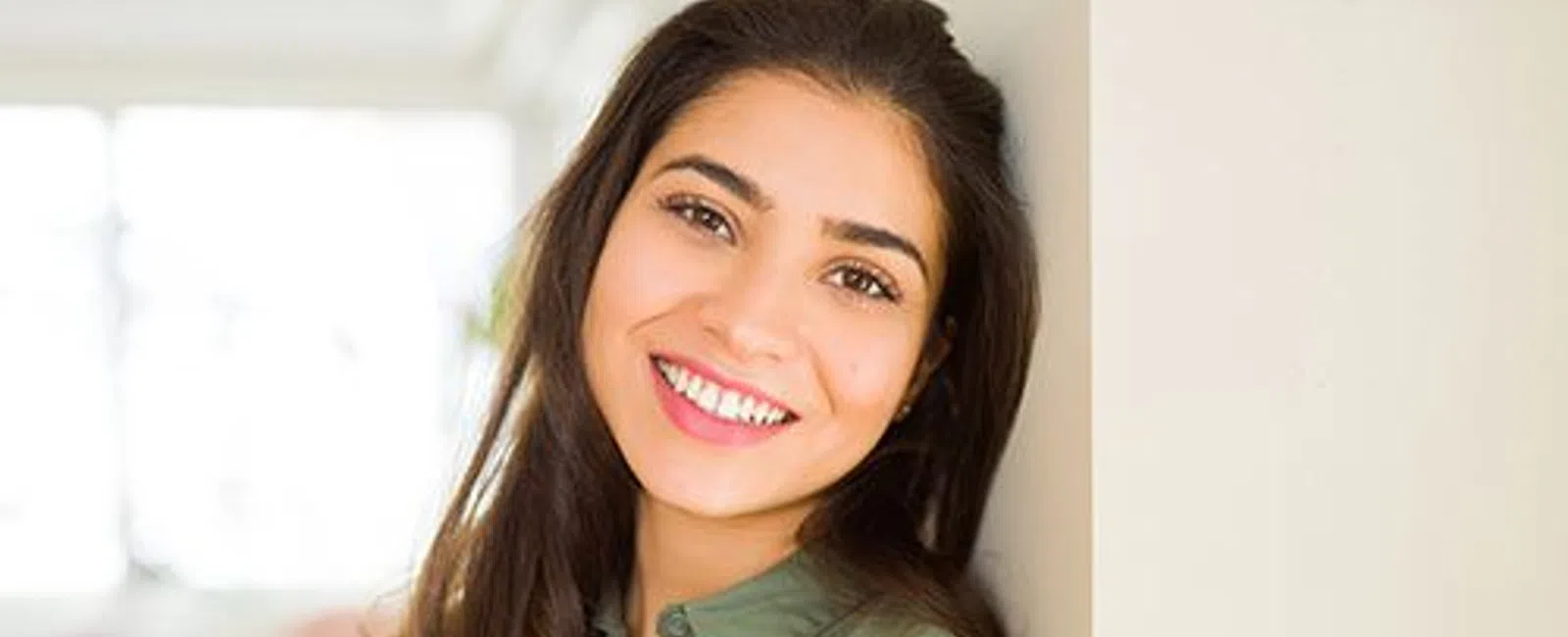 Smiling young woman with long dark hair wearing green shirt leaning against wall indoors.