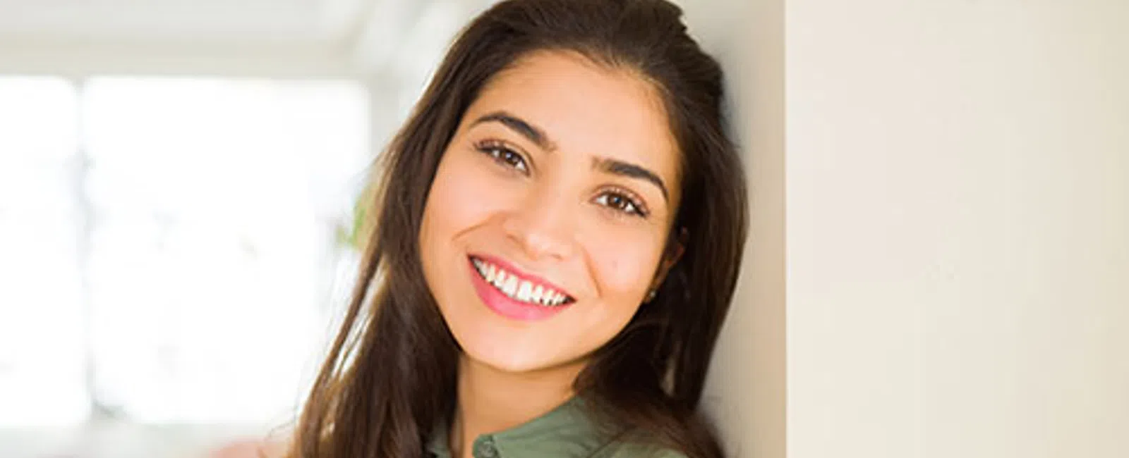 Smiling young woman in green shirt leaning against wall in bright indoor setting.