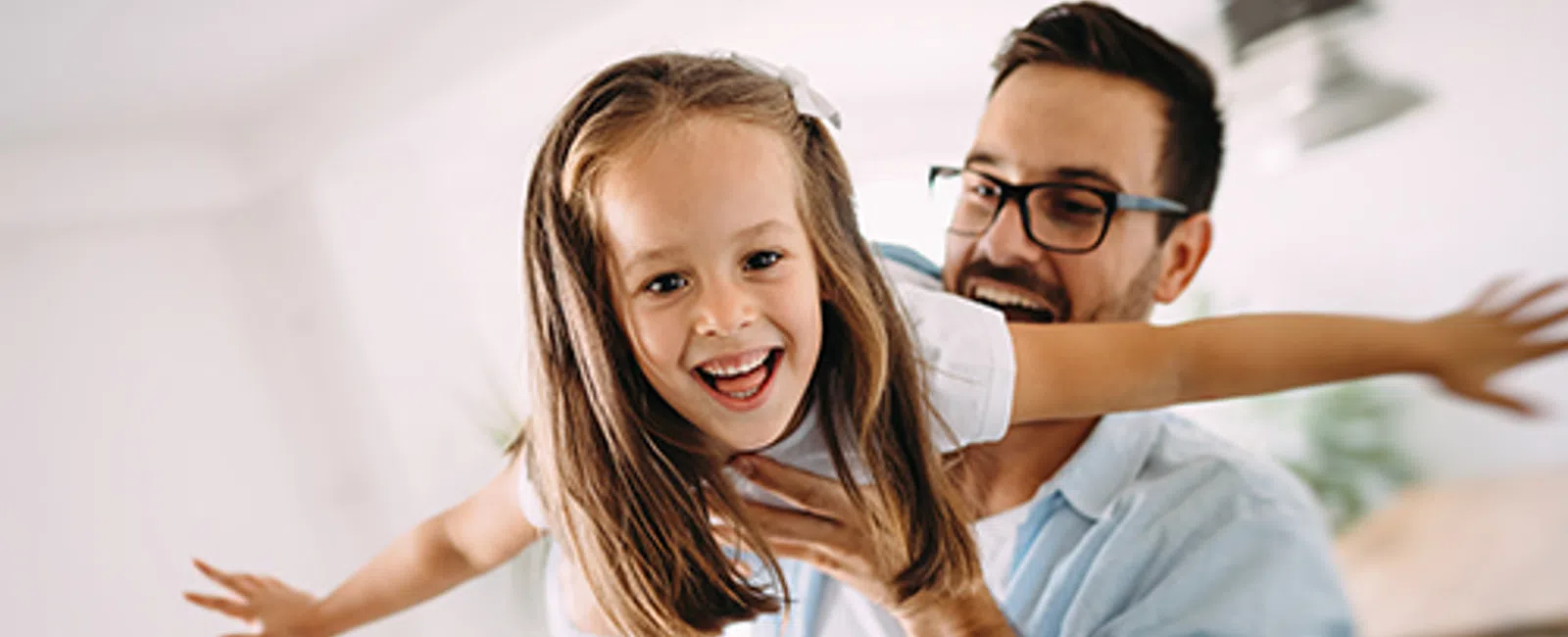 Father playfully lifts smiling daughter with outstretched arms in bright living room.