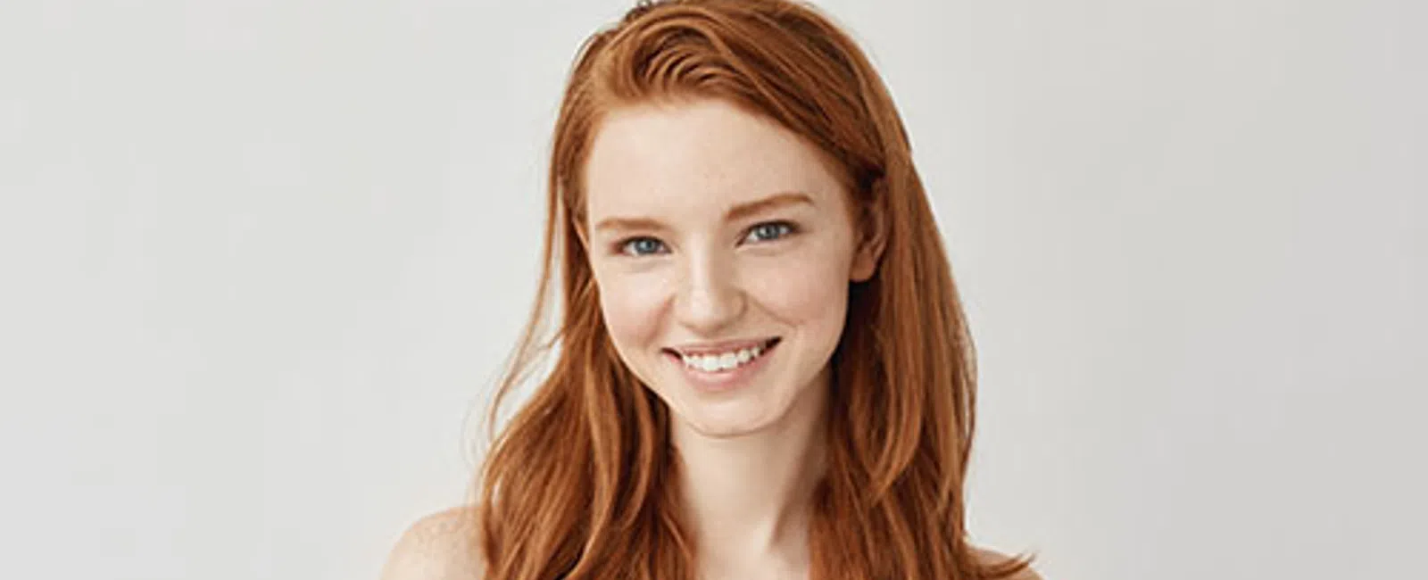 Smiling young woman with long red hair wearing a blue sleeveless top against a plain background.