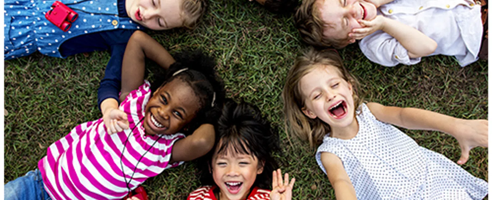 Six diverse children lying on grass in a circle, smiling and laughing joyfully on a sunny day.