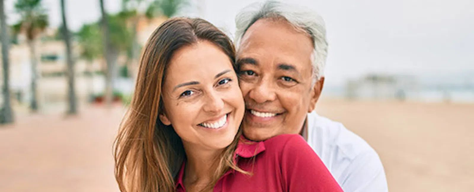 Smiling mature man hugging young woman in red sweater outdoors with palm trees and beach in background