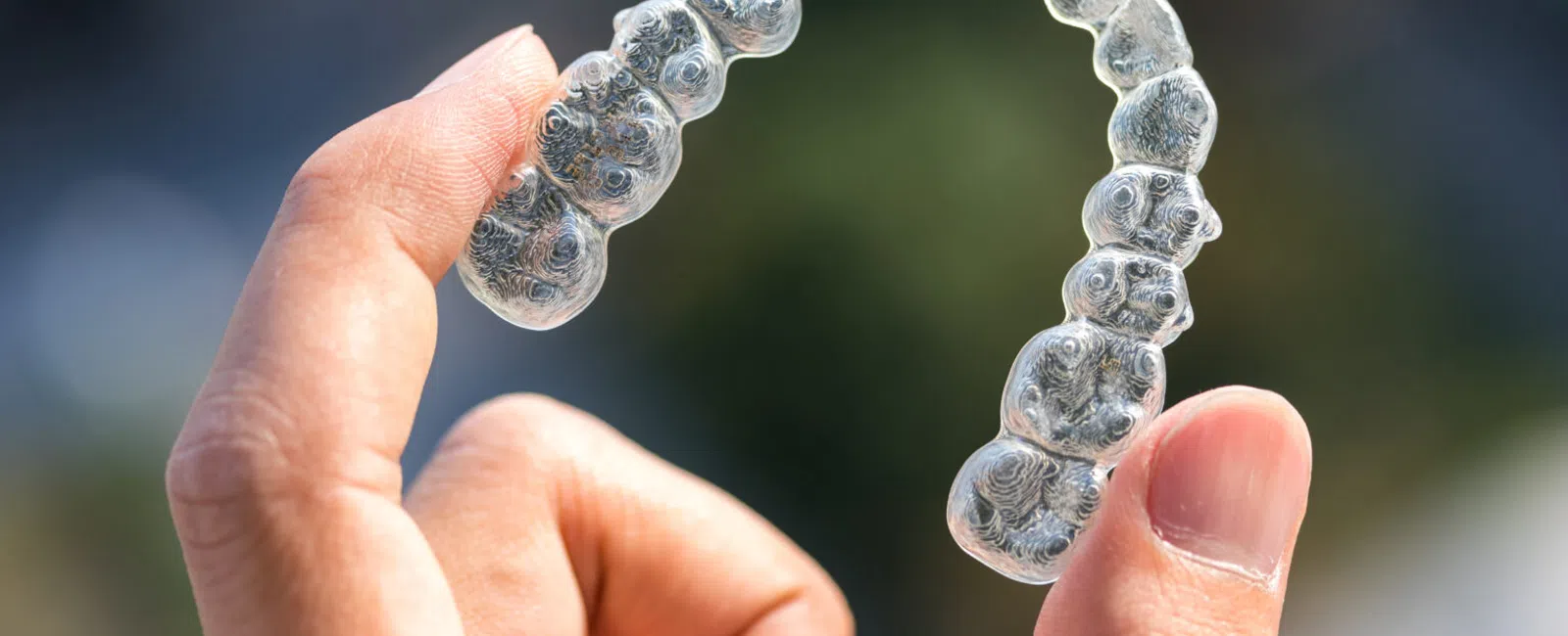 Hand holding a clear dental aligner or retainer against a blurred outdoor background.