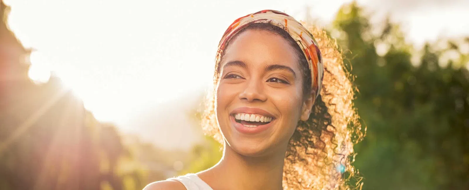 Smiling young woman with curly hair and a headband enjoying sunlight outdoors in a nature setting.