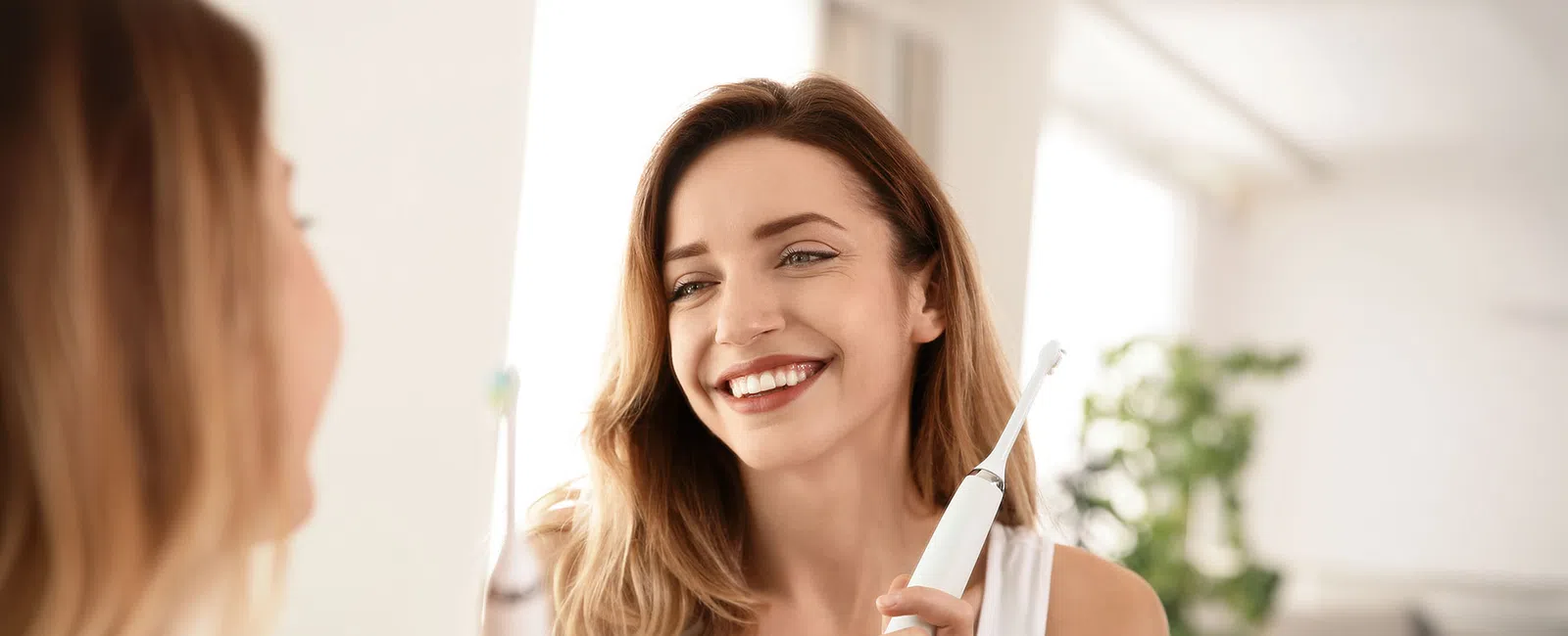 Smiling woman holding electric toothbrush in bathroom mirror, promoting dental hygiene and oral care routine.