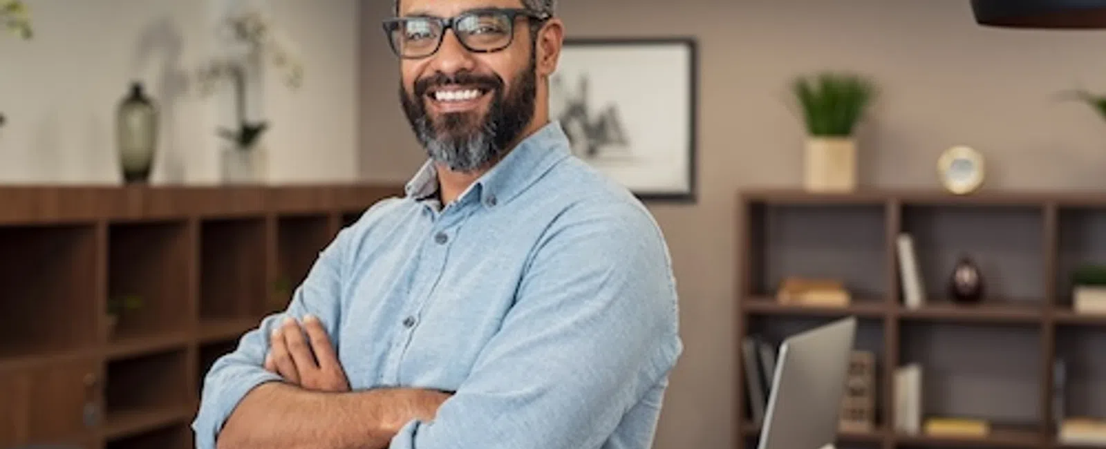 Smiling man with glasses and beard in blue shirt standing confidently in modern office with crossed arms.