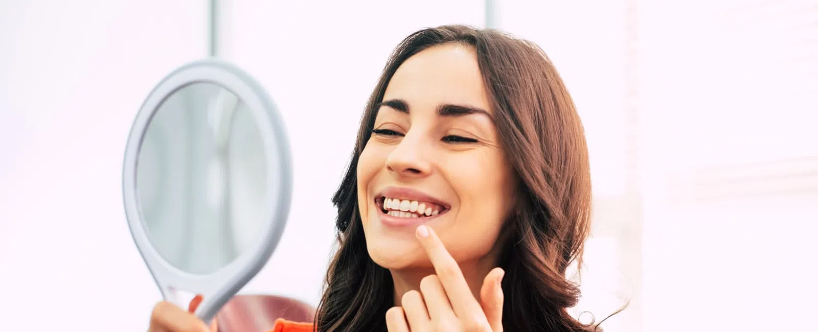 Young woman smiling and inspecting her teeth in a handheld mirror indoors with natural light.