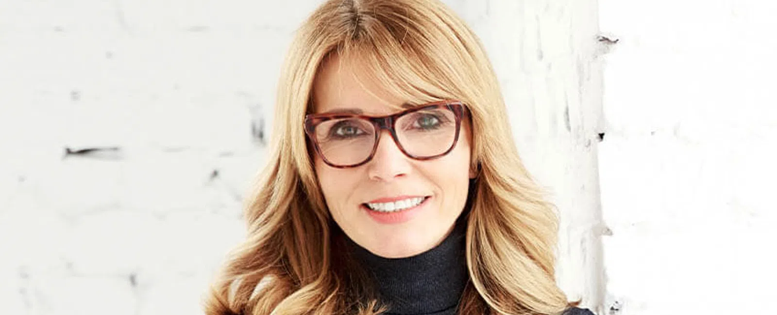 Smiling woman with blonde hair and glasses wearing a dark turtleneck sweater standing against a white brick wall.