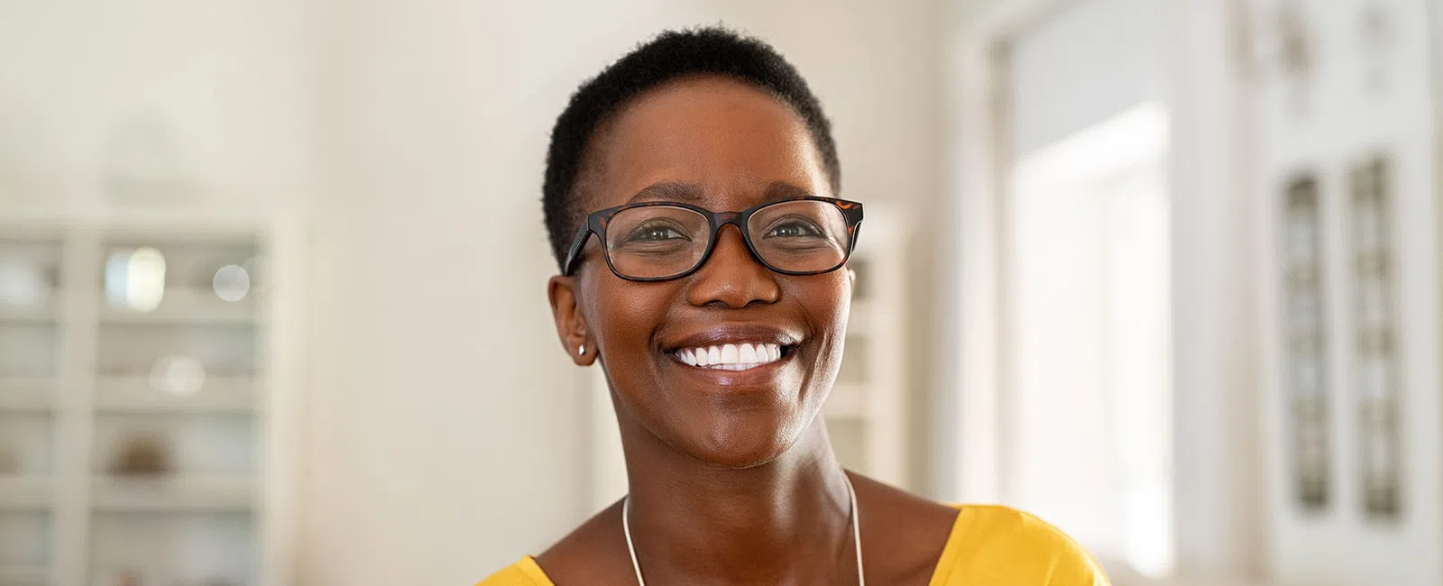 Smiling woman with short hair and glasses wearing a yellow top in a bright, modern room.