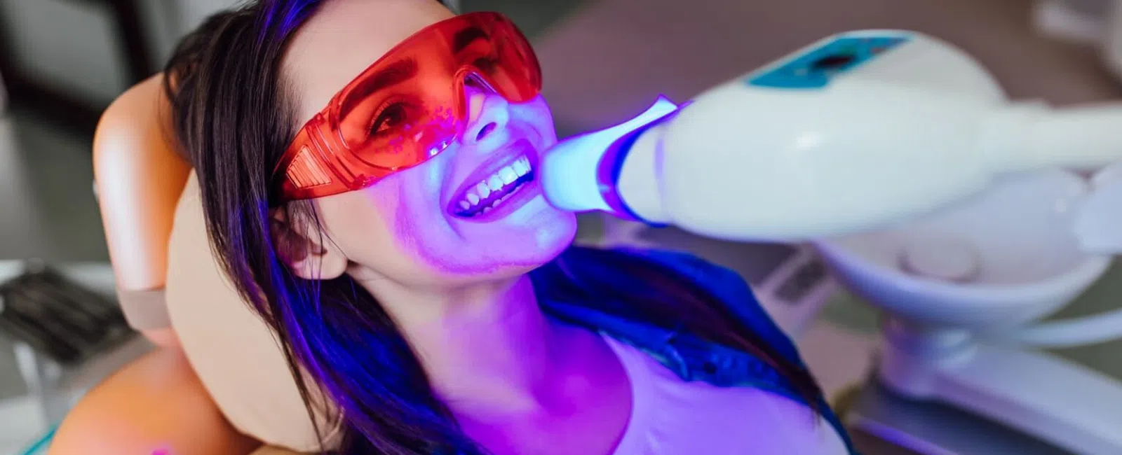 Woman wearing protective red glasses undergoing dental teeth whitening with a bright blue light device.