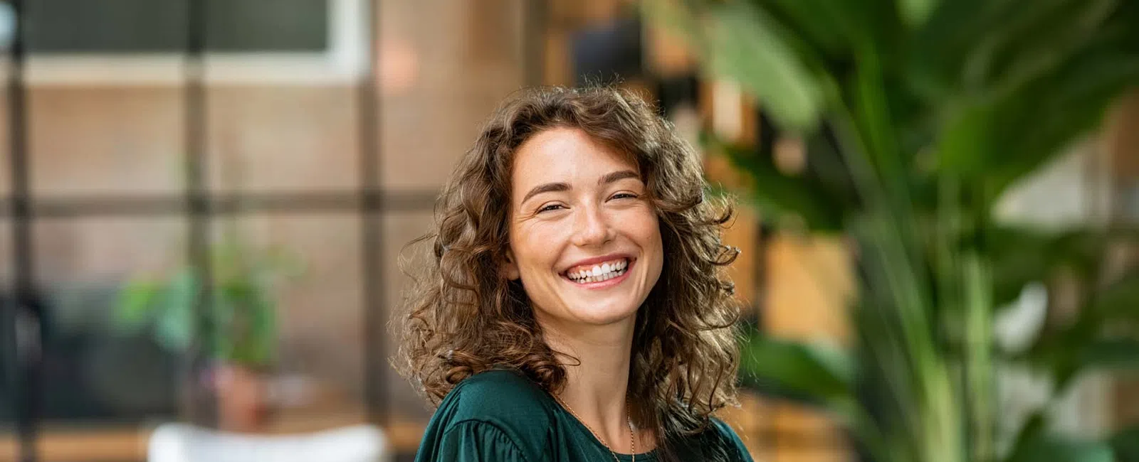 Smiling curly woman in green blouse standing with arms crossed in modern office with plants and blurred background