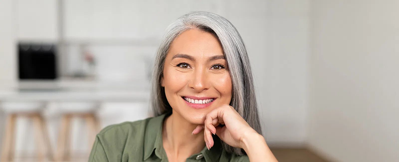 Smiling middle-aged woman with gray hair wearing green shirt in a bright modern kitchen.