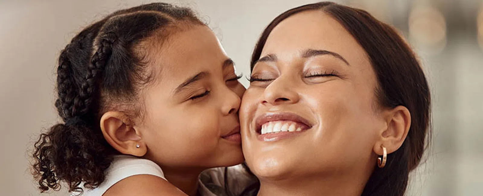 Young girl kisses her smiling mother on the cheek while hugging her tightly in a warm family moment.