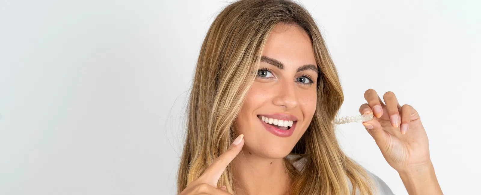Smiling woman with blonde hair holds a clear dental aligner near her mouth wearing a white shirt and smartwatch.