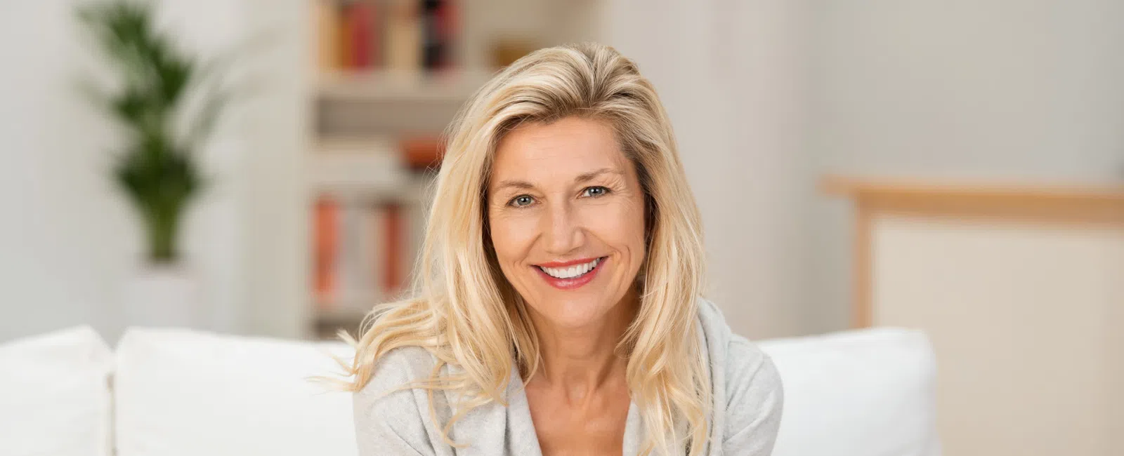 Smiling blonde woman sitting on white couch in a bright, modern living room with bookshelves and plants.