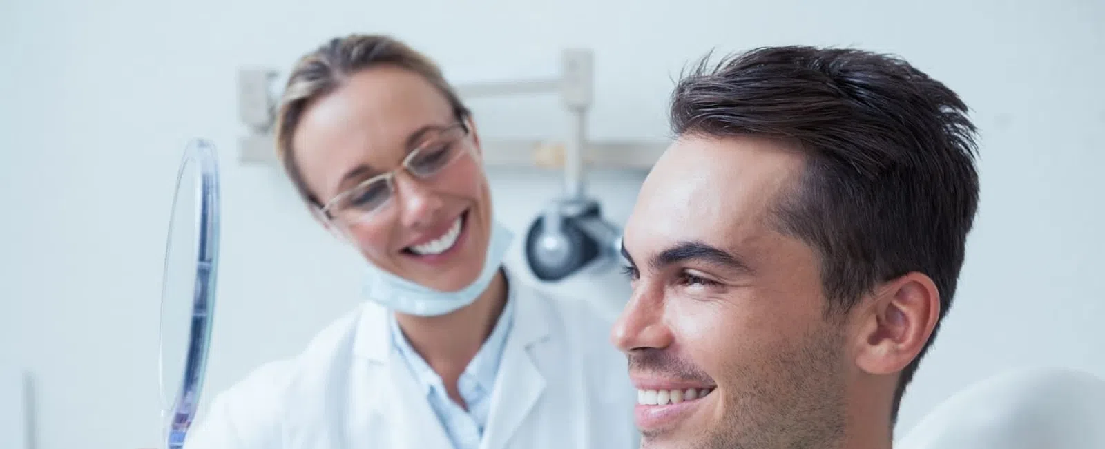 Smiling man checks teeth in handheld mirror while dentist observes in bright dental office
