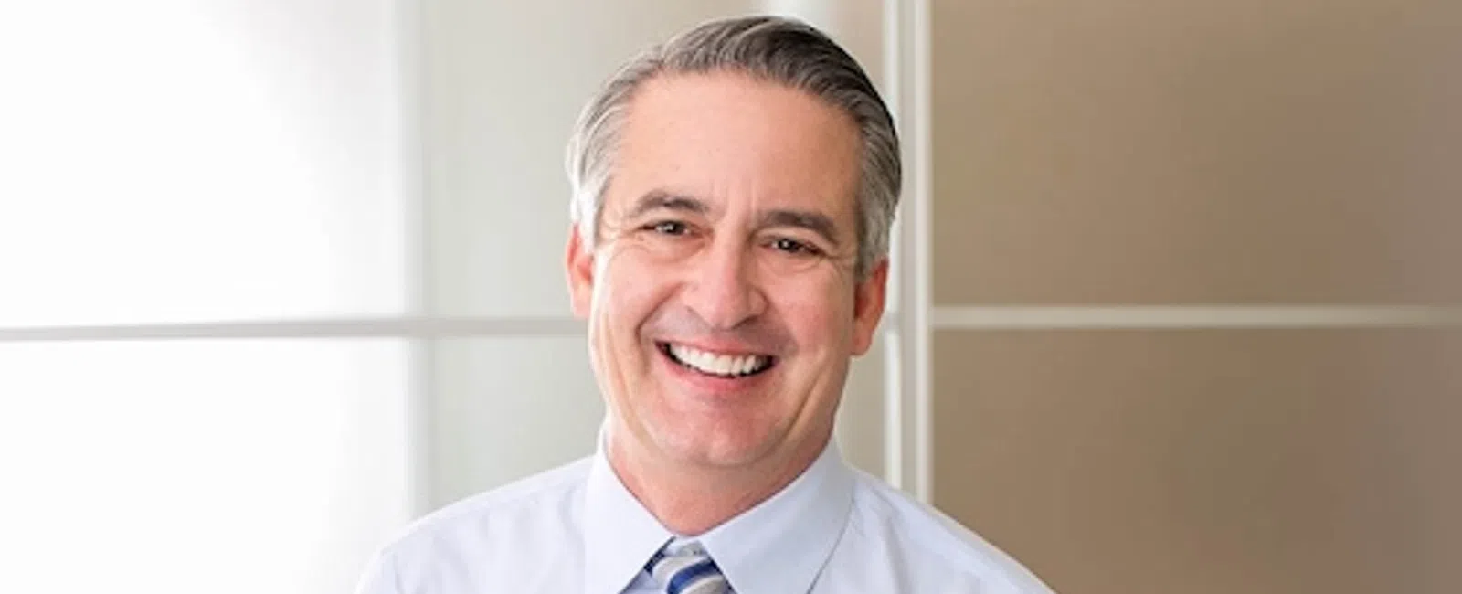 Smiling middle-aged man in light blue shirt and striped tie standing against a neutral office background.