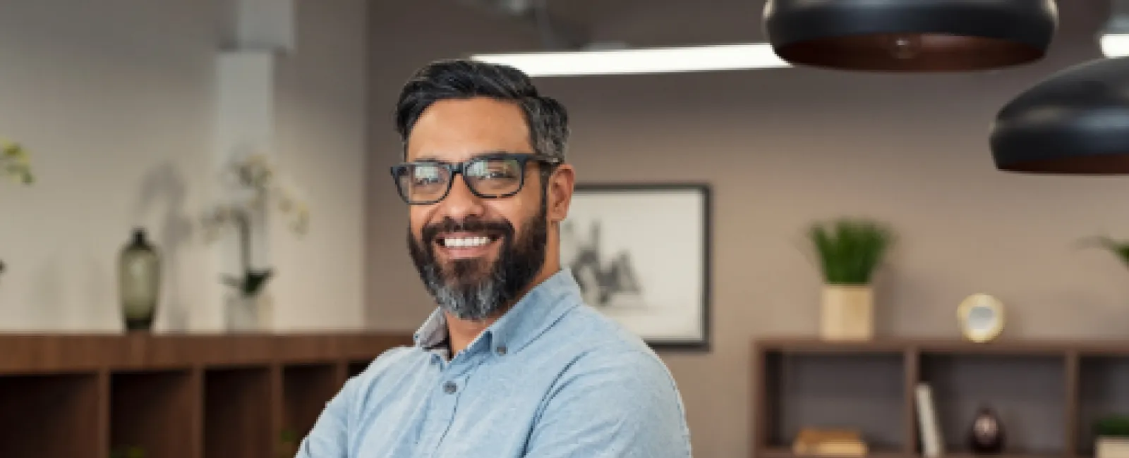 Smiling man with glasses and beard standing confidently in modern office with crossed arms