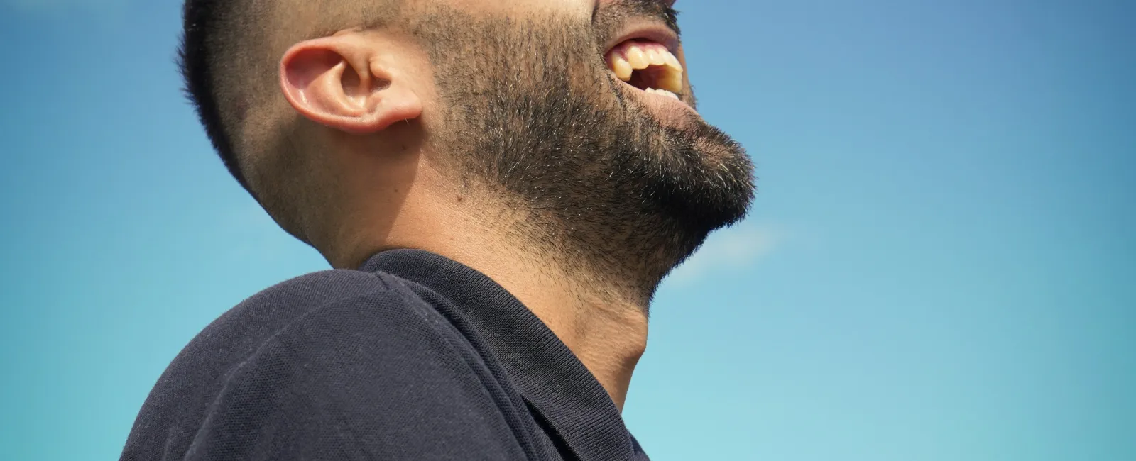 Side view of a bearded man laughing joyfully against a clear blue sky with a few clouds.