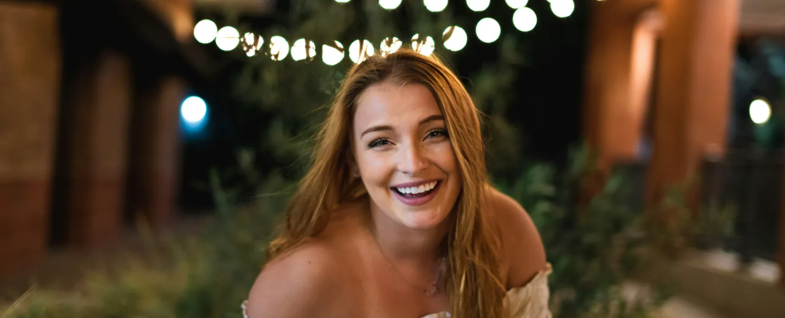 Smiling woman with long hair wearing an off-shoulder top outdoors at night with string lights in the background