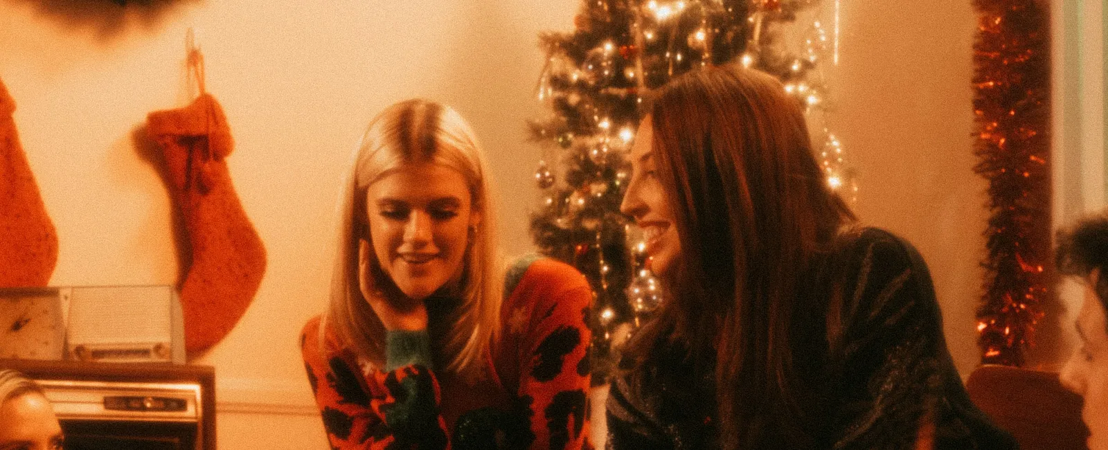 Friends exchanging Christmas gifts by a decorated tree in a cozy, warmly lit living room with festive decorations.