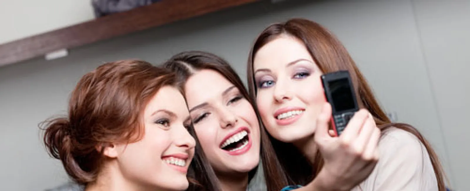 Three women smiling and taking a selfie with a flip phone indoors, capturing a happy moment together.