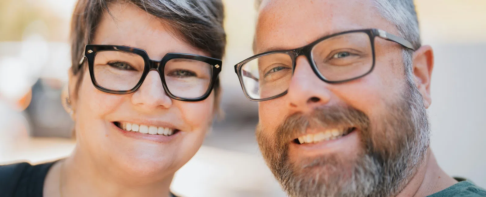 Smiling middle-aged couple wearing glasses taking a close-up selfie outdoors in daylight.